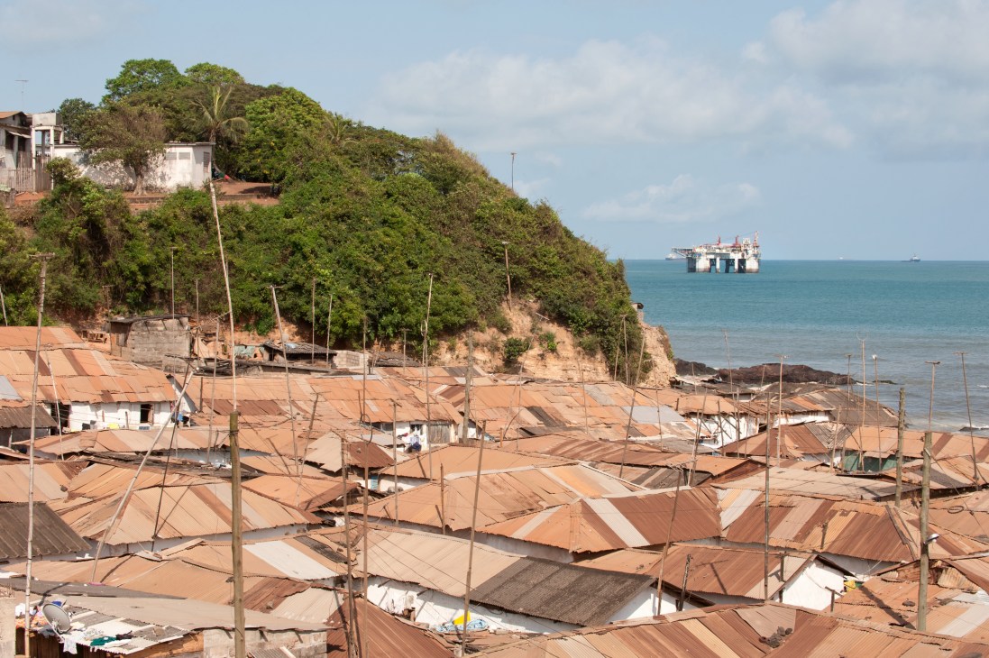 Fishing village rooftops overlooking ocean with offshore oil rig visible on the horizon.