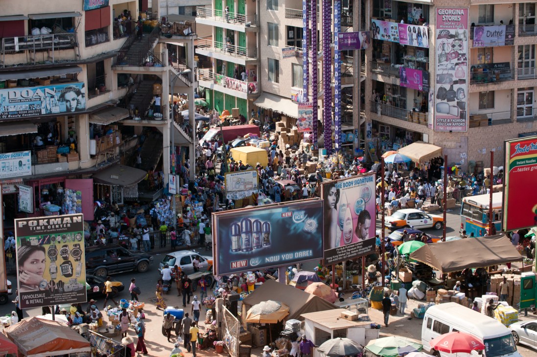 Aerial view of Makola market in central Accra showing crowded streets, market stalls, and colorful advertising billboards on surrounding buildings.