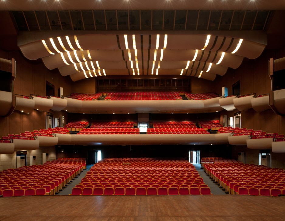 Interior of a modern theatre auditorium with three tiers of red seats and curved architectural lighting fixtures on the ceiling.
