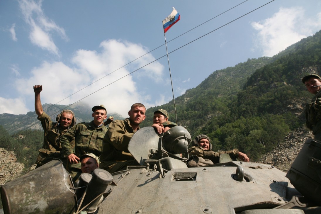 Russian soldiers in military uniforms aboard a tank with a Russian flag flying, mountainous terrain in background.