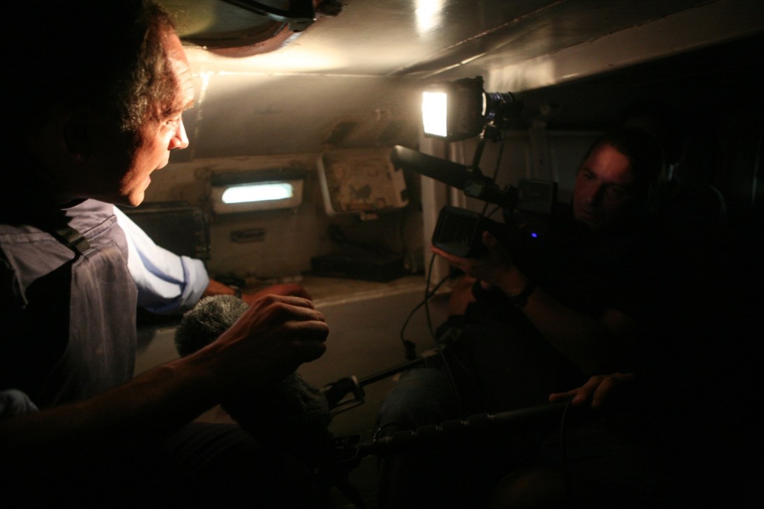 Journalist reporting inside a Russian armoured personnel carrier with camera operator and lighting equipment in dim conditions.