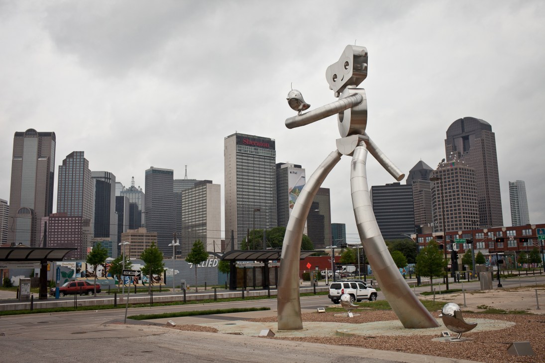 'Traveling Man' public sculpture at Deep Ellum's DART light rail station with Dallas skyline behind.