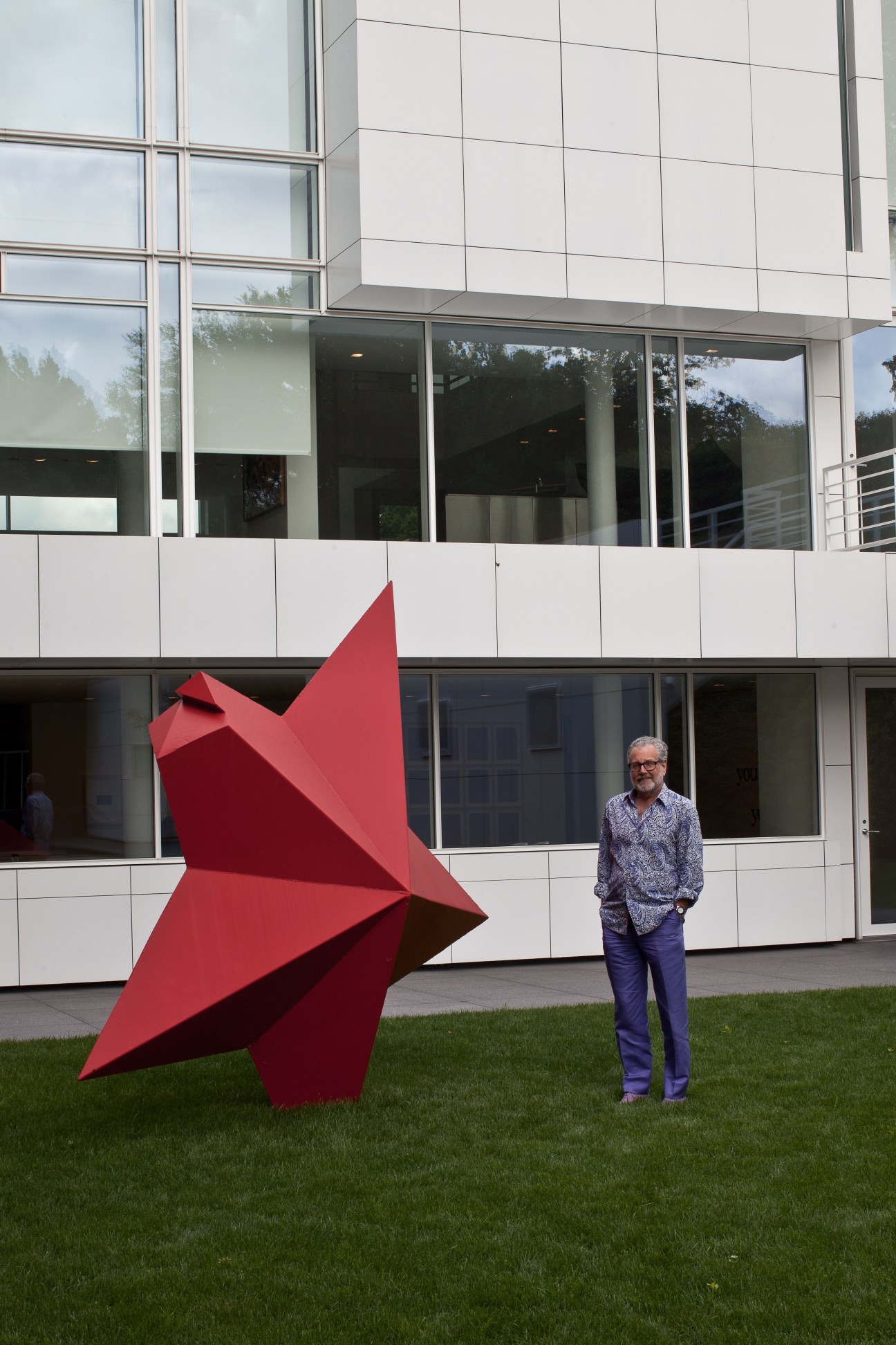 Howard Rachofsky stands beside a large red abstract sculpture on the lawn of a modern white building.