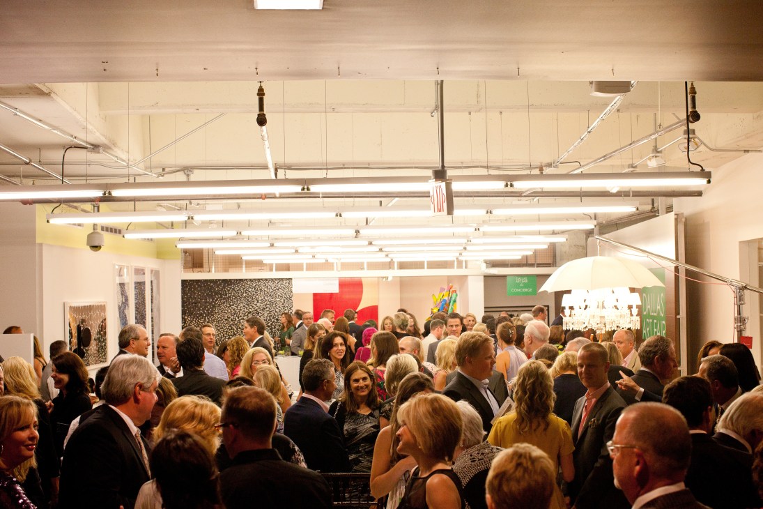 Crowds of visitors in business attire mingle at the Dallas Art Fair opening gala in a modern gallery space with colorful artworks displayed on walls.