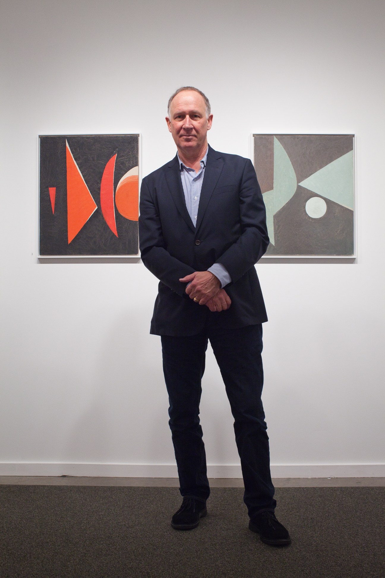 Dallas gallery owner Barry Whistler standing in front of two abstract artworks with geometric shapes in red and green.