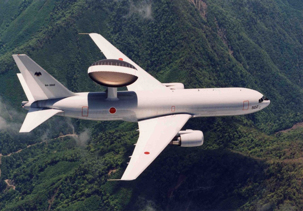 Japanese military surveillance aircraft with distinctive radar dome flying over forested mountains.