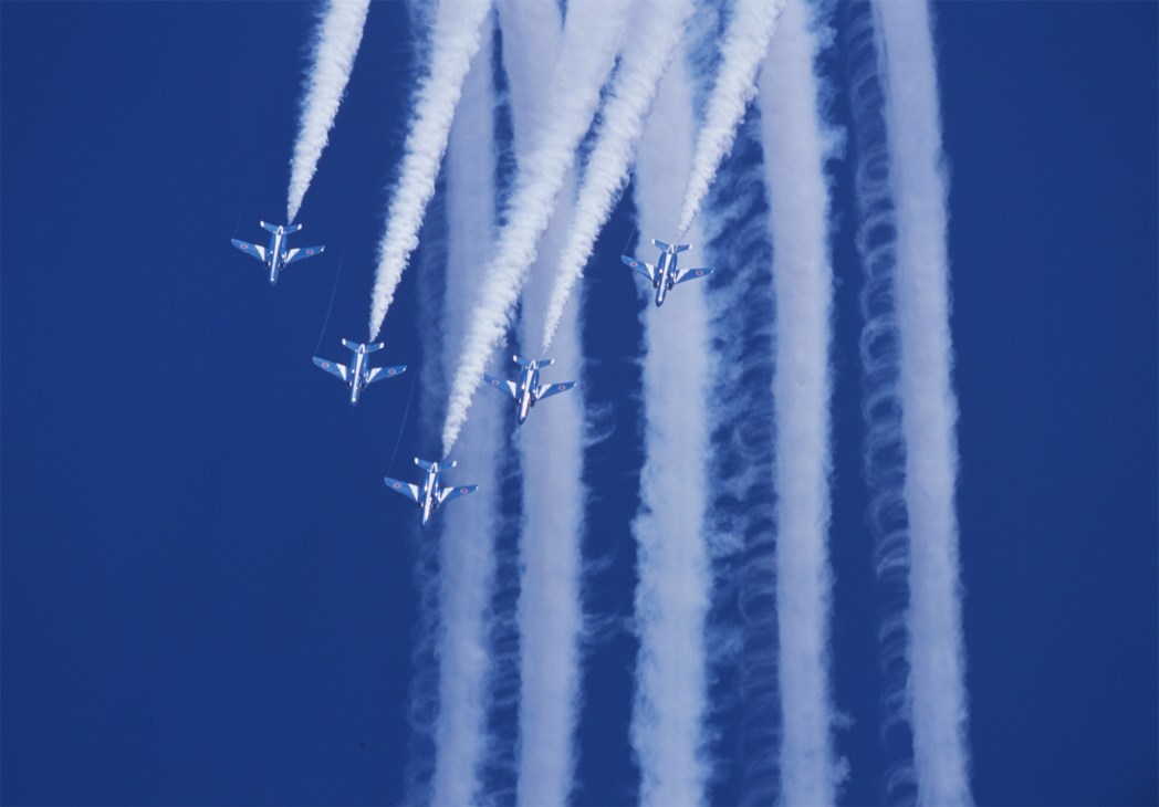 Five military fighter jets in formation flying against a blue sky with white smoke trails streaming behind them.