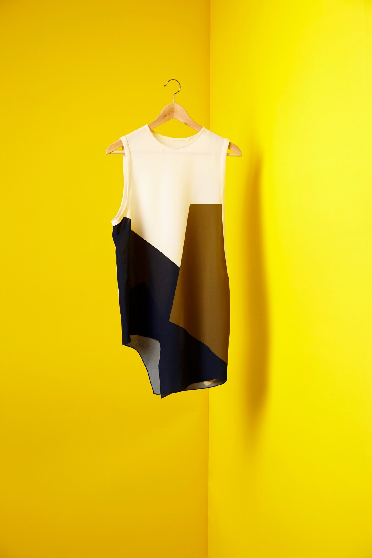 Sleeveless dress with cream, black, and olive block colour panels hanging on wooden hanger against yellow background.