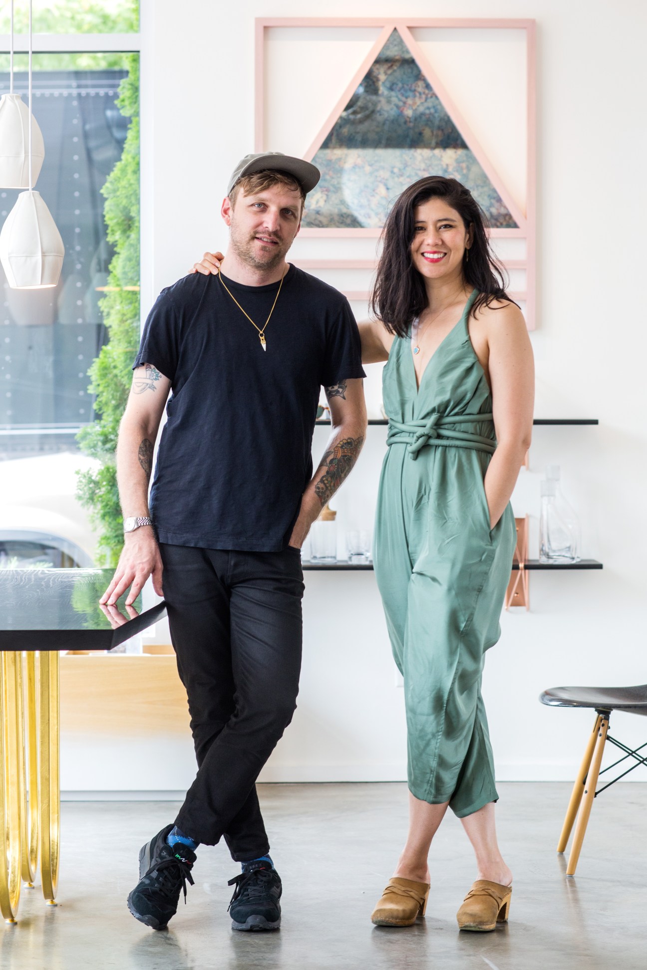 Two people standing in a bright interior space with framed artwork and hanging plants.
