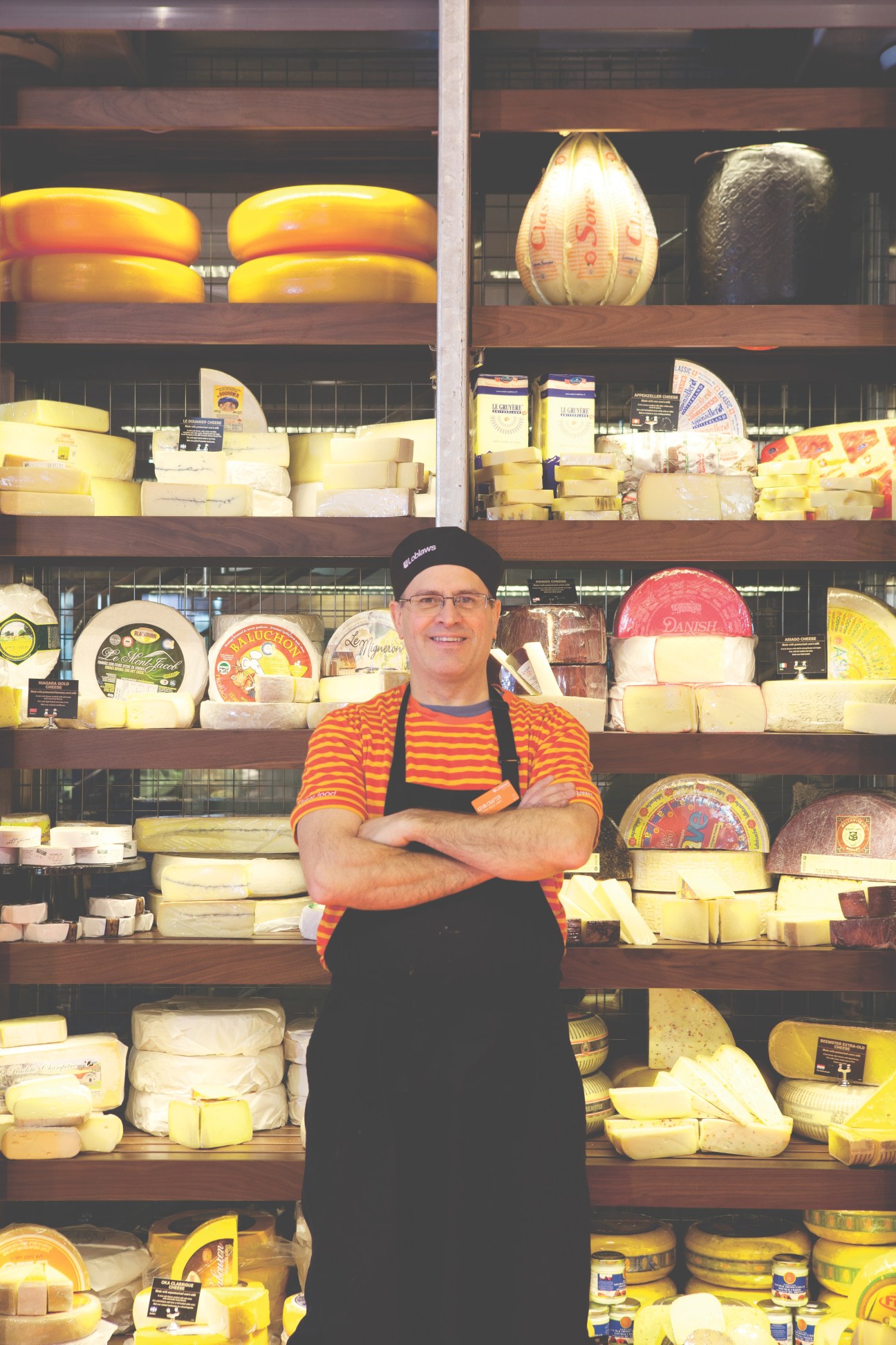 Cheese counter staff member in striped shirt standing before towering display of over 400 cheese varieties.