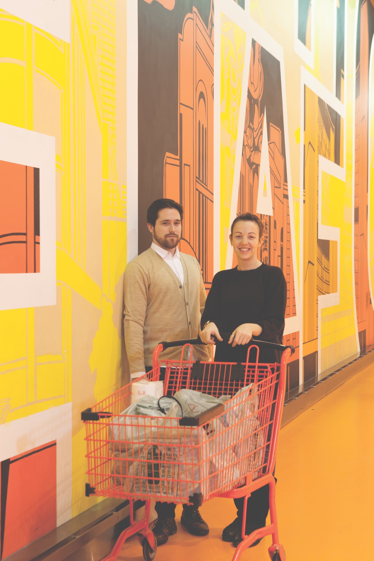 Two customers with a red shopping cart stand in front of yellow and orange mural artwork inside Loblaws supermarket.