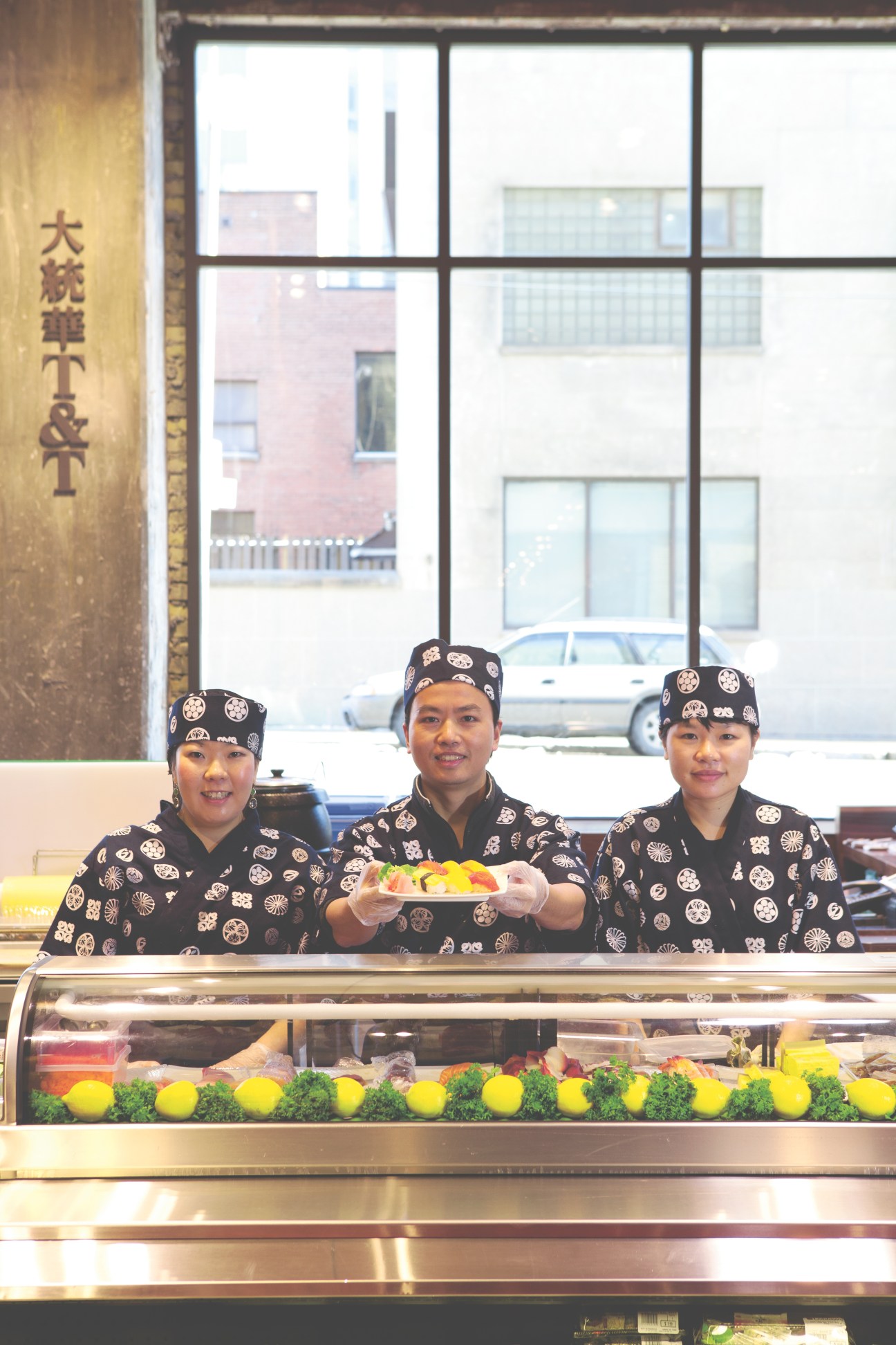 Three sushi chefs in matching black uniforms with white circular patterns serve sushi at the T&T Supermarket counter.