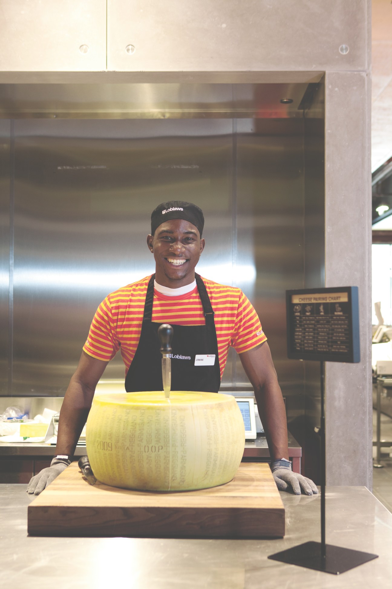 Loblaws employee in striped shirt and apron smiles beside a large wheel of Parmesan cheese at the counter.