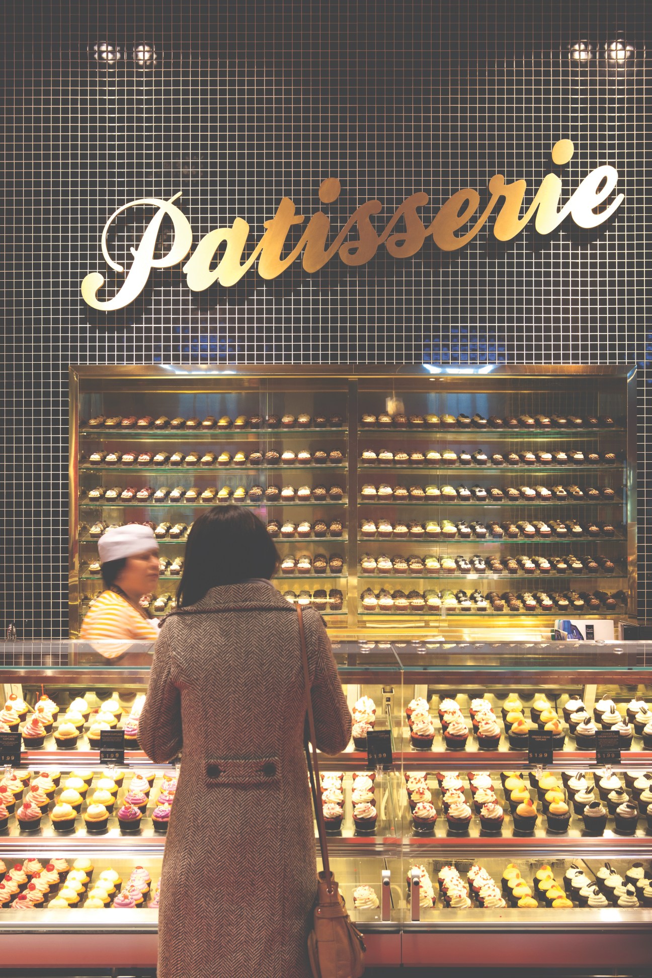 Customer viewing illuminated display cases of cupcakes at Ace Bakery shop-in-shop counter with signage.