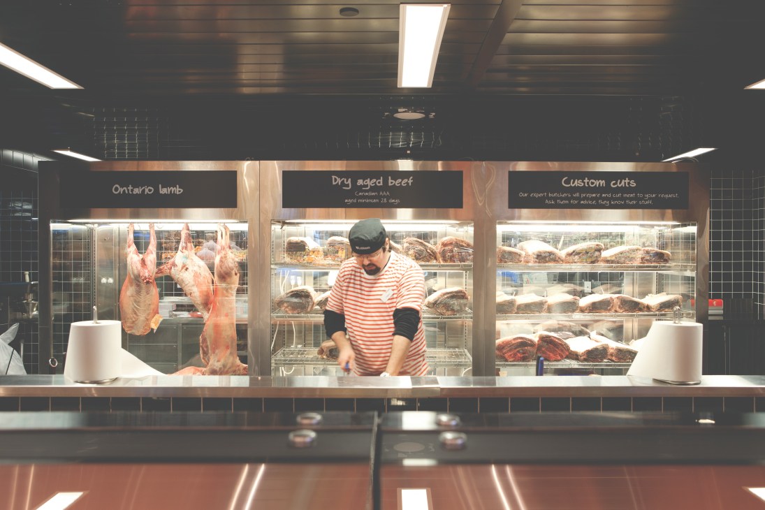 Butcher in striped shirt working at meat counter with hanging lamb and packaged cuts displayed behind glass.
