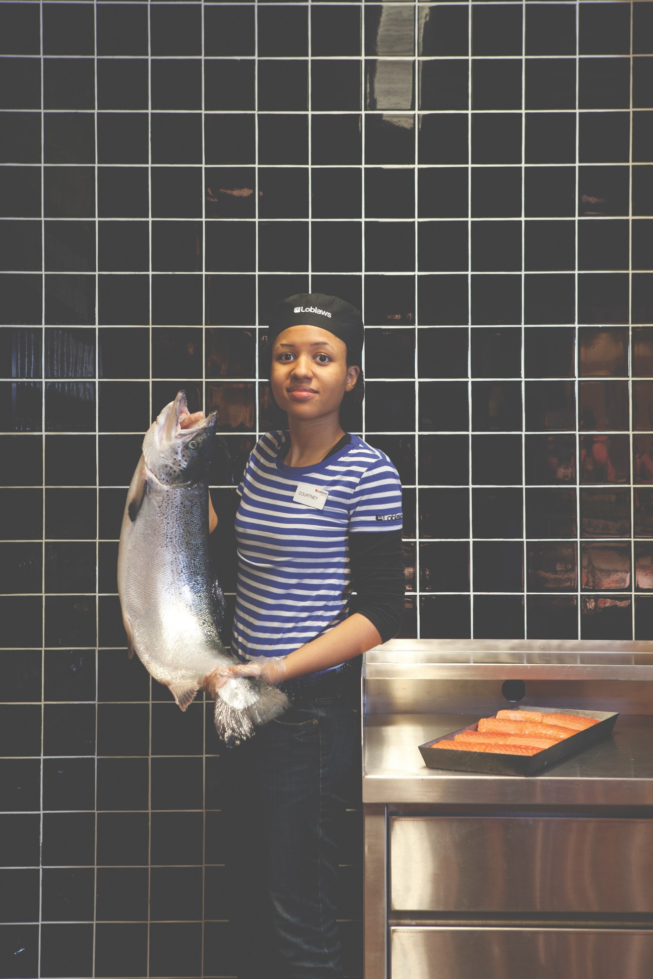 Loblaws employee in striped shirt and cap holding a fresh salmon at the fishmonger counter.