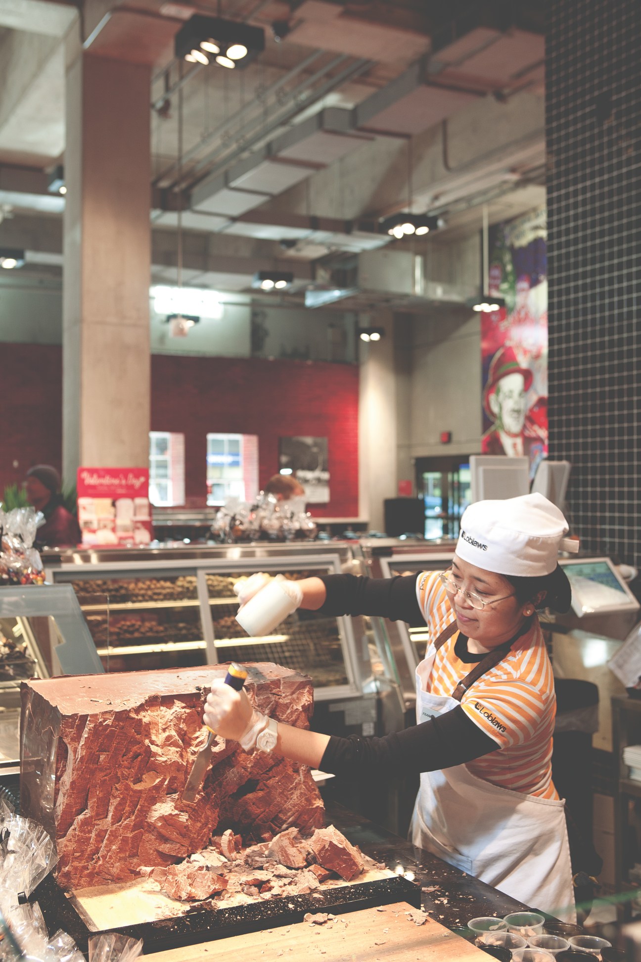 Loblaws employee in striped shirt and white cap chipping chocolate from a large block at the store counter.