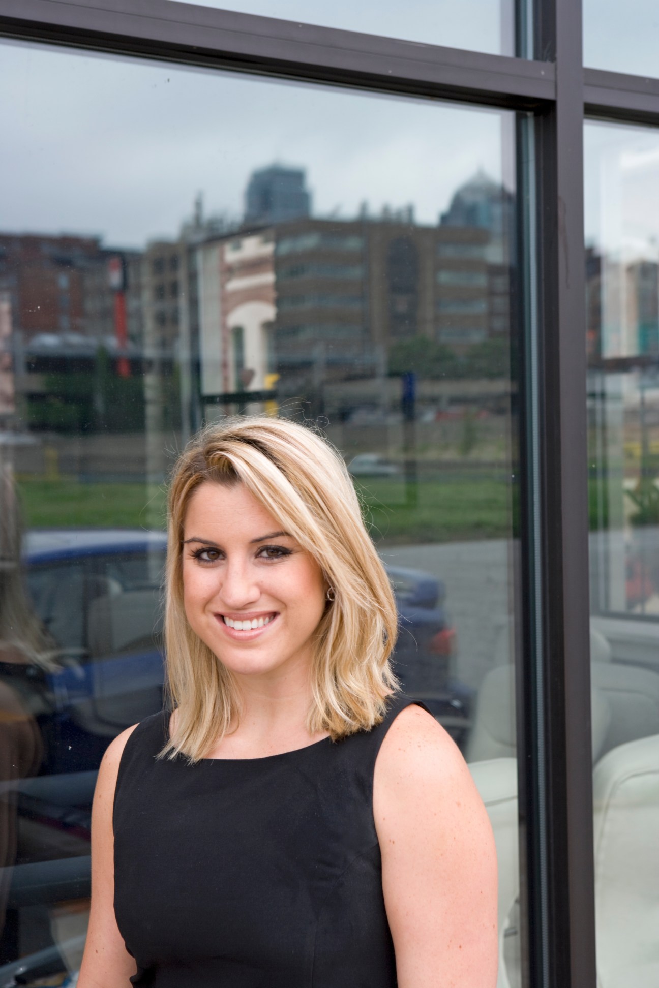 Woman smiling at camera in black sleeveless top by window with Kansas City skyline behind.