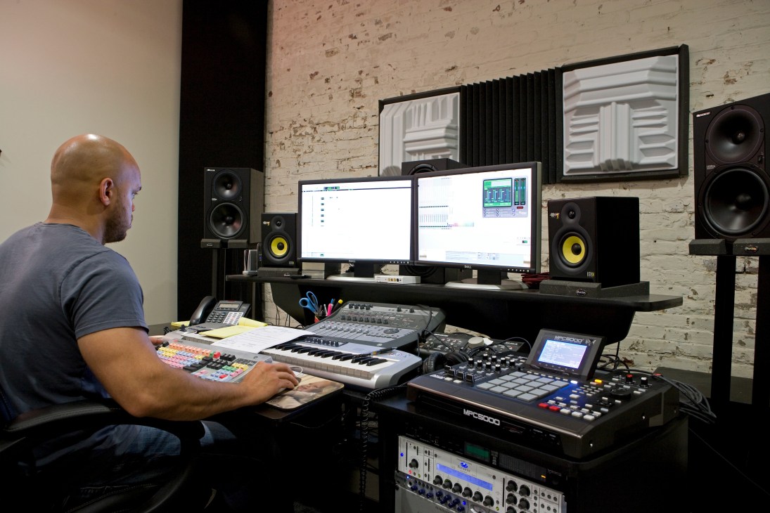 Man working at a music production desk with keyboards, monitors, and studio speakers in a brick-walled sound studio.