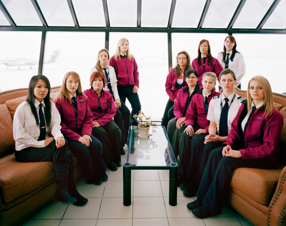 Female pilot students in maroon jackets sitting in an airport lounge with aircraft visible outside.