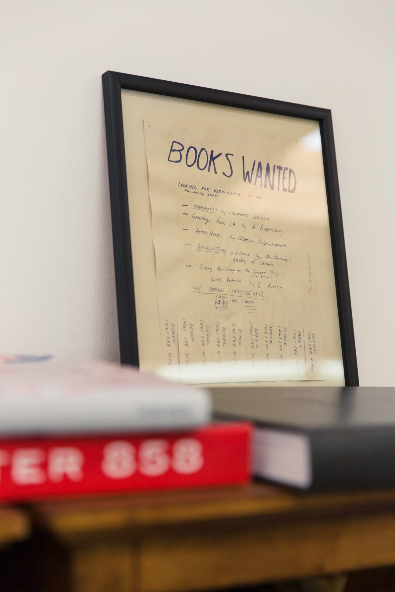 Framed artwork by Derek Sullivan displaying a handwritten "Books Wanted" notice with book titles listed.
