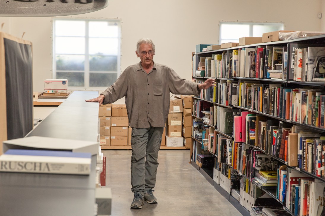 Artist Ed Ruscha stands in his library surrounded by organized shelves of books and cardboard storage boxes.
