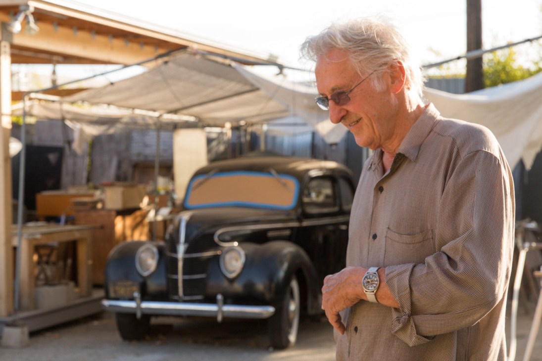 Ed Ruscha stands in the courtyard of his Culver City studio, wearing a light pink shirt and glasses, with a vintage black car behind him under canvas shade structures.