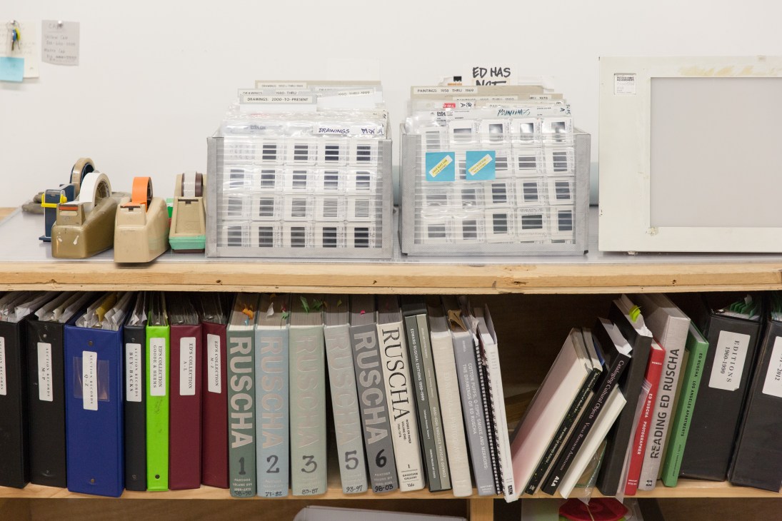 Organized shelf in Ed Ruscha's studio office with tape dispensers, storage drawers, and reference books.