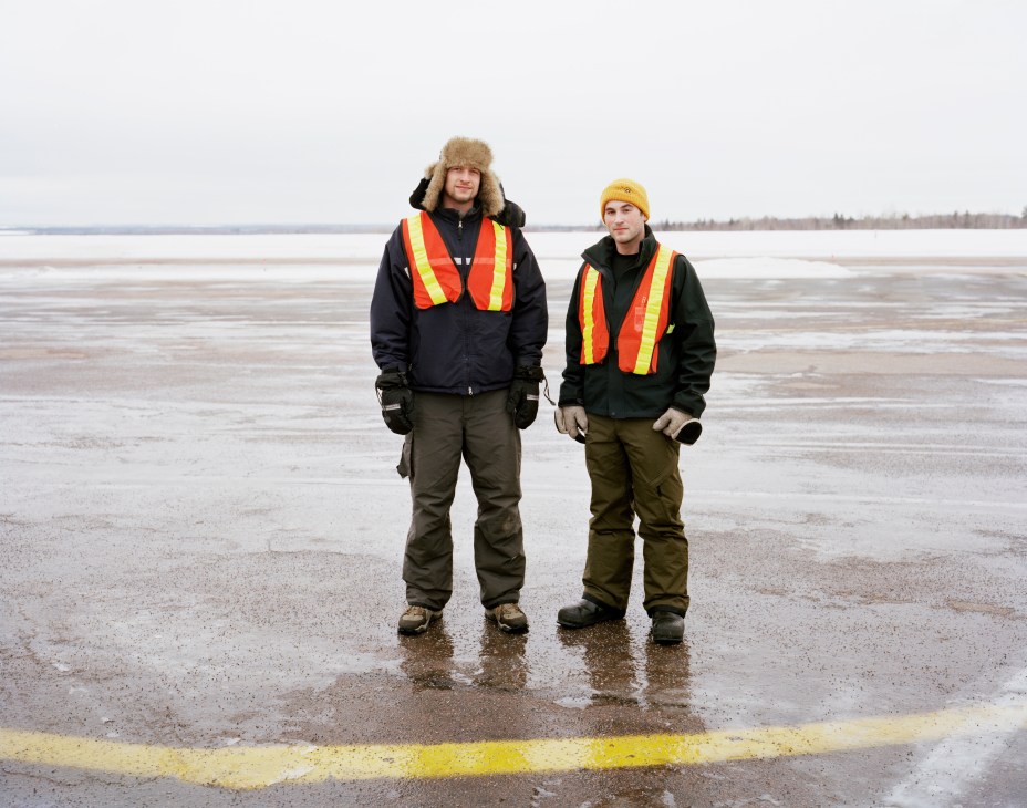 Two flight students in safety vests stand on a snow-covered runway beside a frozen lake.