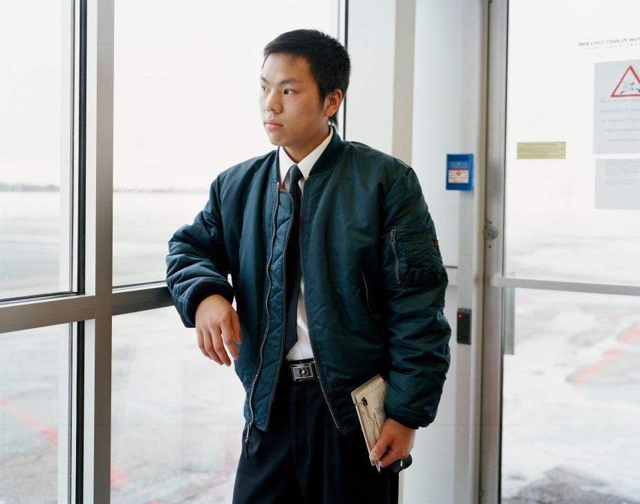 Chinese pilot student Li Tao standing by a window overlooking an airfield, wearing a dark blue jacket.
