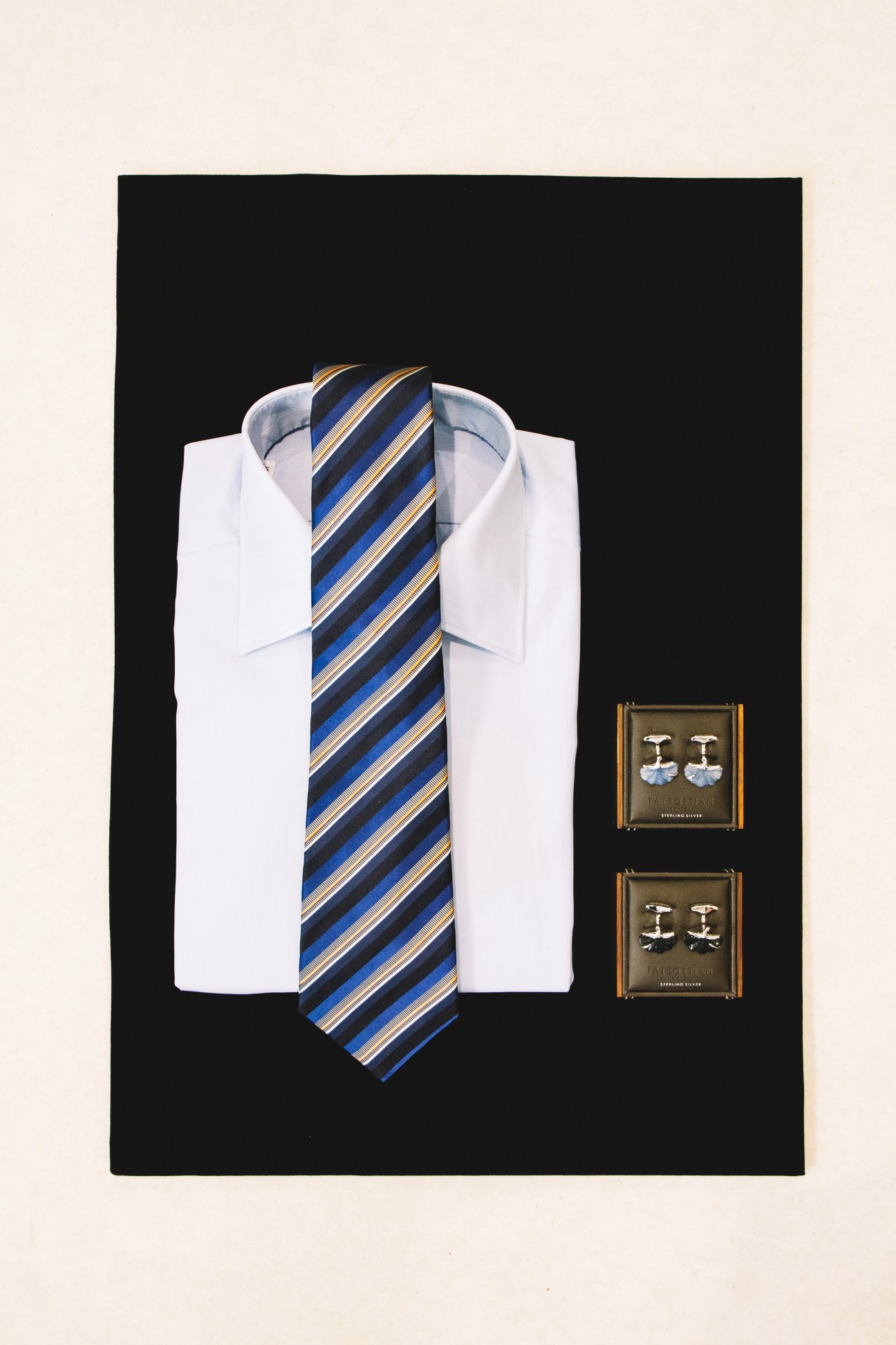 White dress shirt with blue and gold striped tie and cufflinks arranged on black background.