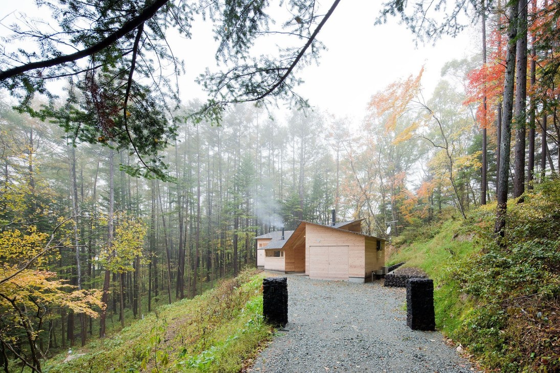 Modern wooden cottage with pitched roof nestled among autumn forest trees with smoke rising from chimney.