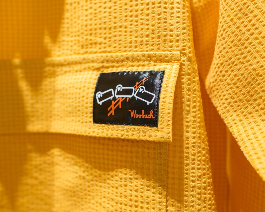 Close-up of black Woolrich Outdoor tag with white logo and orange text on yellow textured fabric