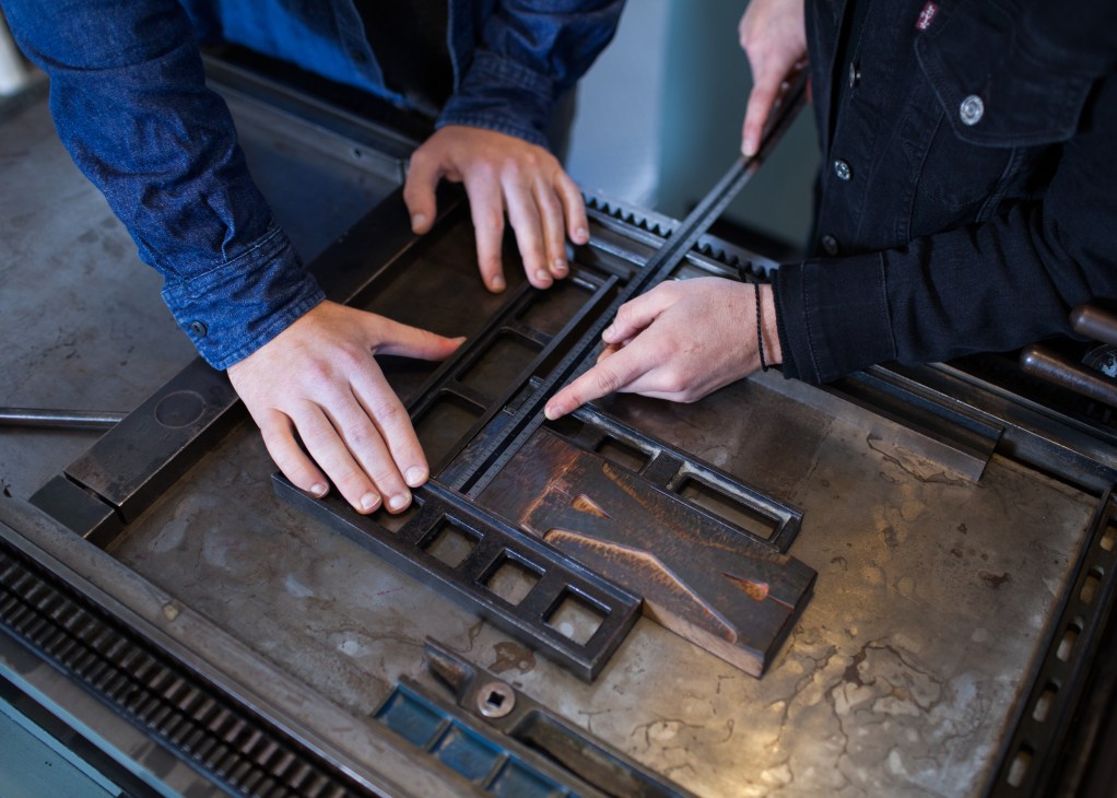 Two hands positioning metal type in a letterpress machine during the typesetting process.