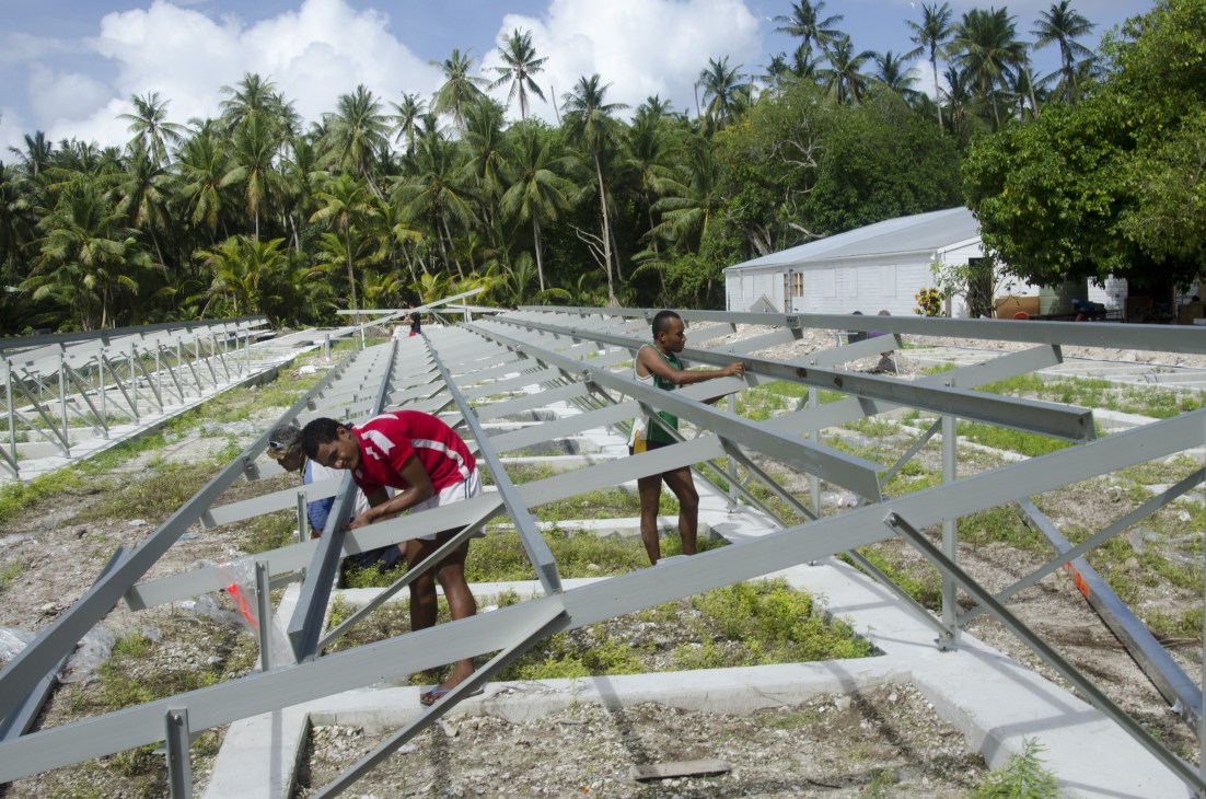Workers install solar panel frames on Tokelau with palm trees and a white building in the background.