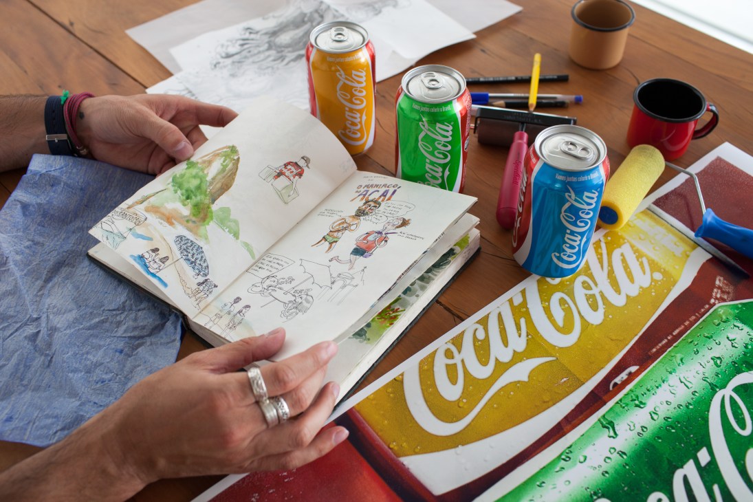 Coca-Cola cans in Brazil's flag colors with sketches and campaign materials on a wooden desk.