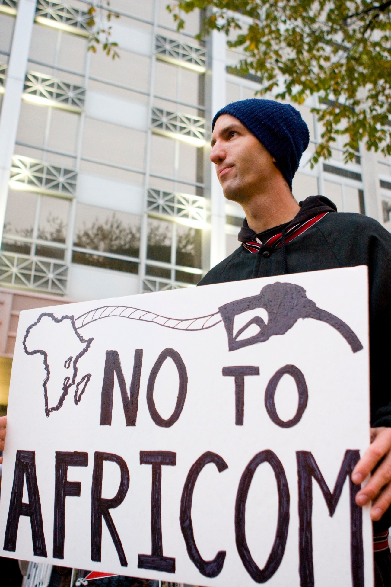 Protester holding sign reading 'NO TO AFRICOM' with Africa map outline outside conference building.