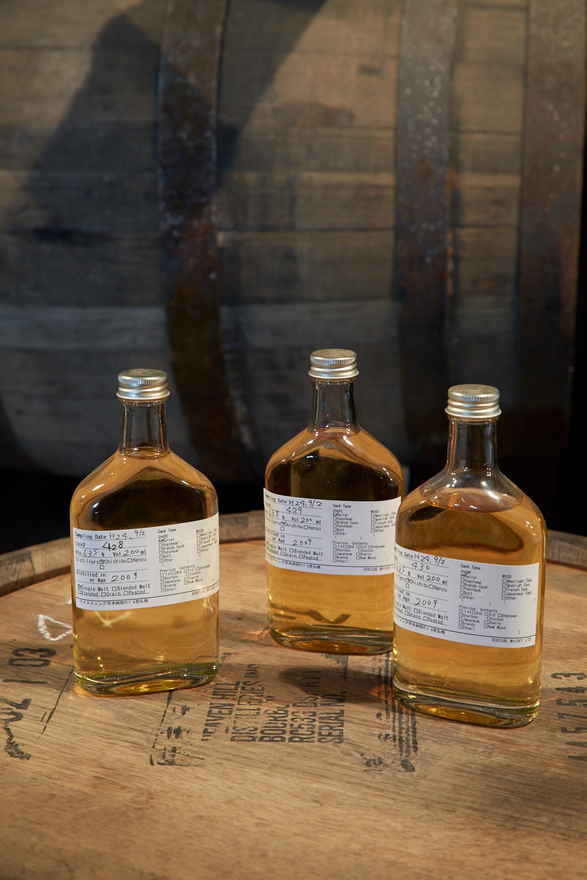 Three whisky sample bottles with detailed labels on wooden surface in front of wooden barrels.