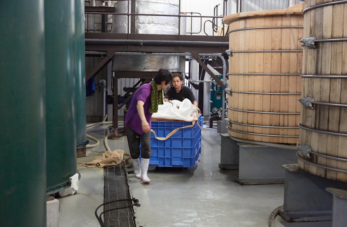 Two workers push a blue crate through a distillery facility lined with large wooden fermentation vessels.