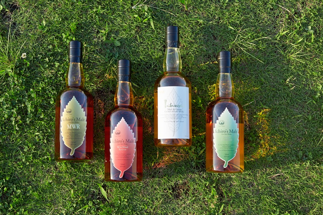 Four bottles of Ichiro's Malt whisky with colorful leaf-shaped labels arranged on green grass.