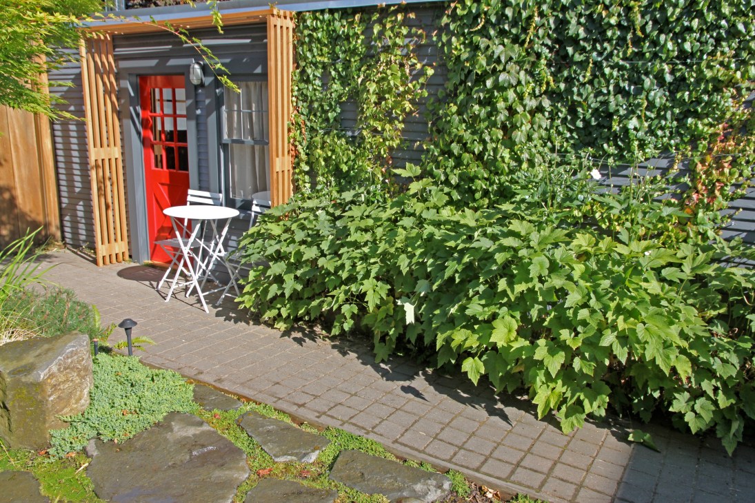 Exterior of Yakuza Garden Cottage with red door, wooden lattice panels, climbing ivy, and white bistro table in lush Portland garden.
