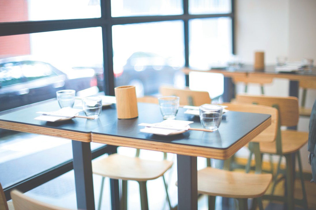 Modern restaurant dining room with wooden chairs, dark tables, and large windows overlooking a city street.