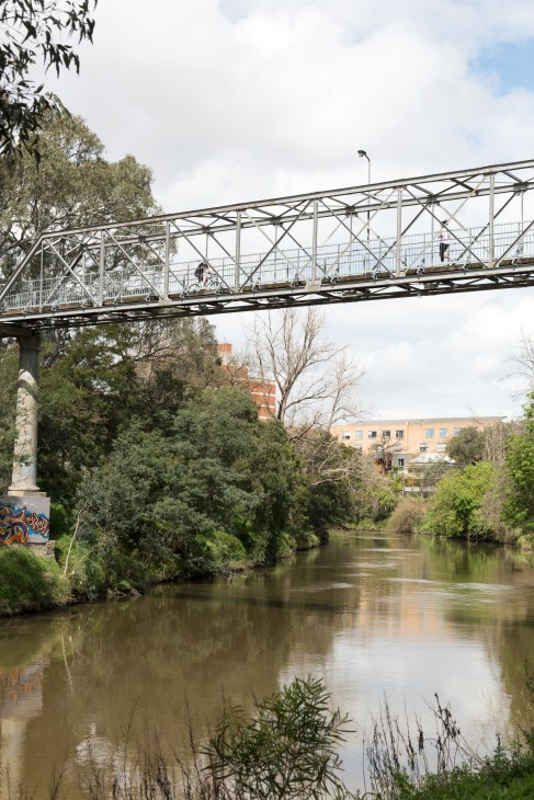 Metal pedestrian bridge spanning the Yarra River with trees and apartment buildings visible in the background.