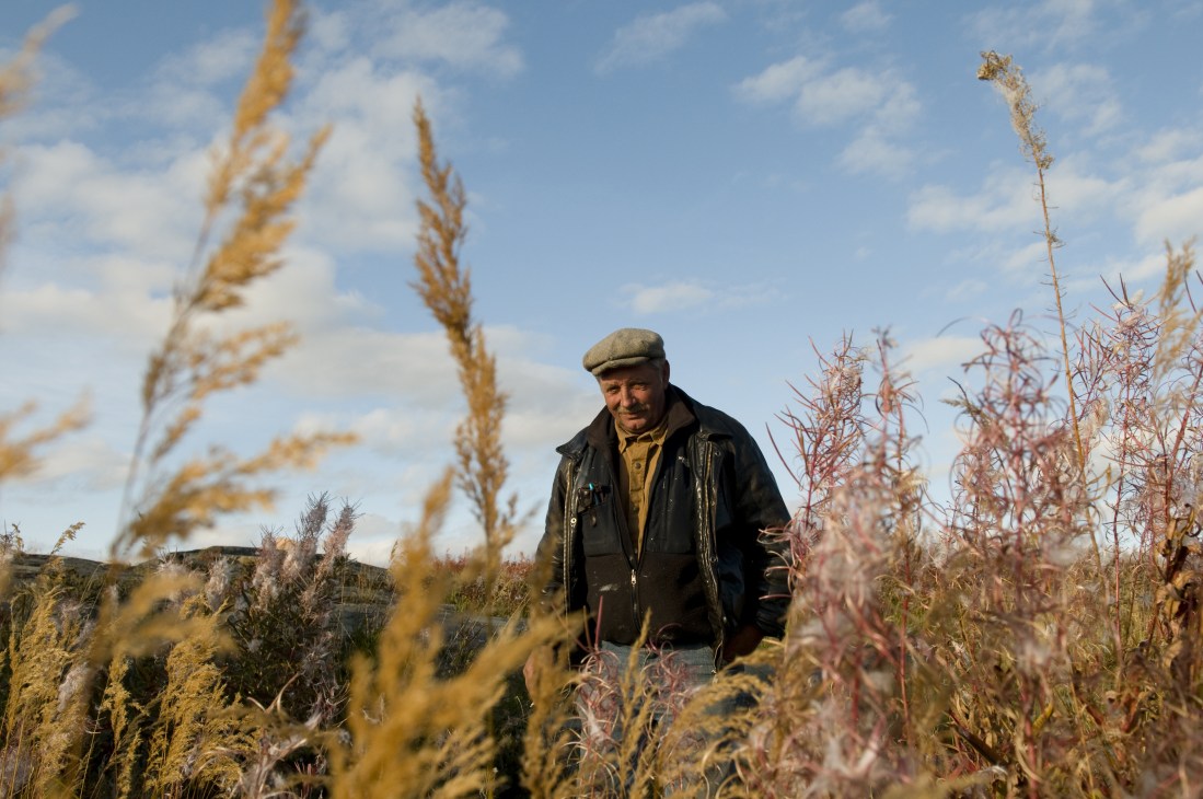 Gary McDonald, a houseboat owner, stands in sparse northern vegetation wearing a flat cap and dark jacket.