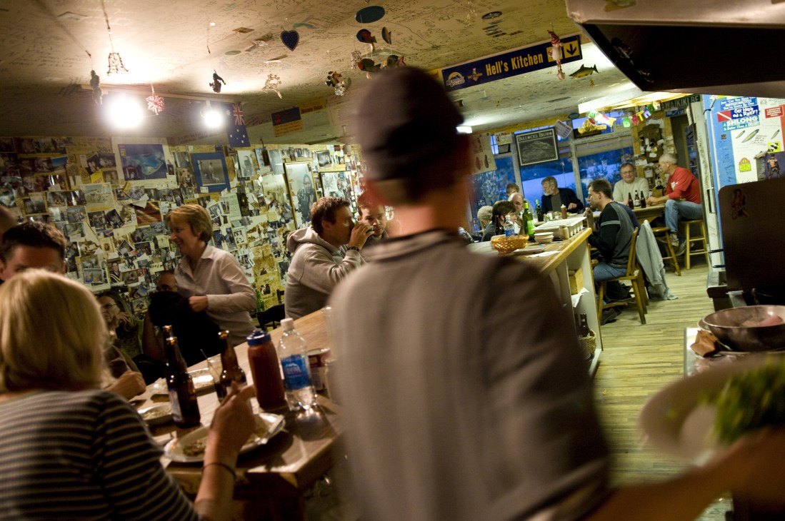 Interior of a frontier-style bar with patrons seated at the counter and standing, walls decorated with memorabilia and signs including Hell's Kitchen.