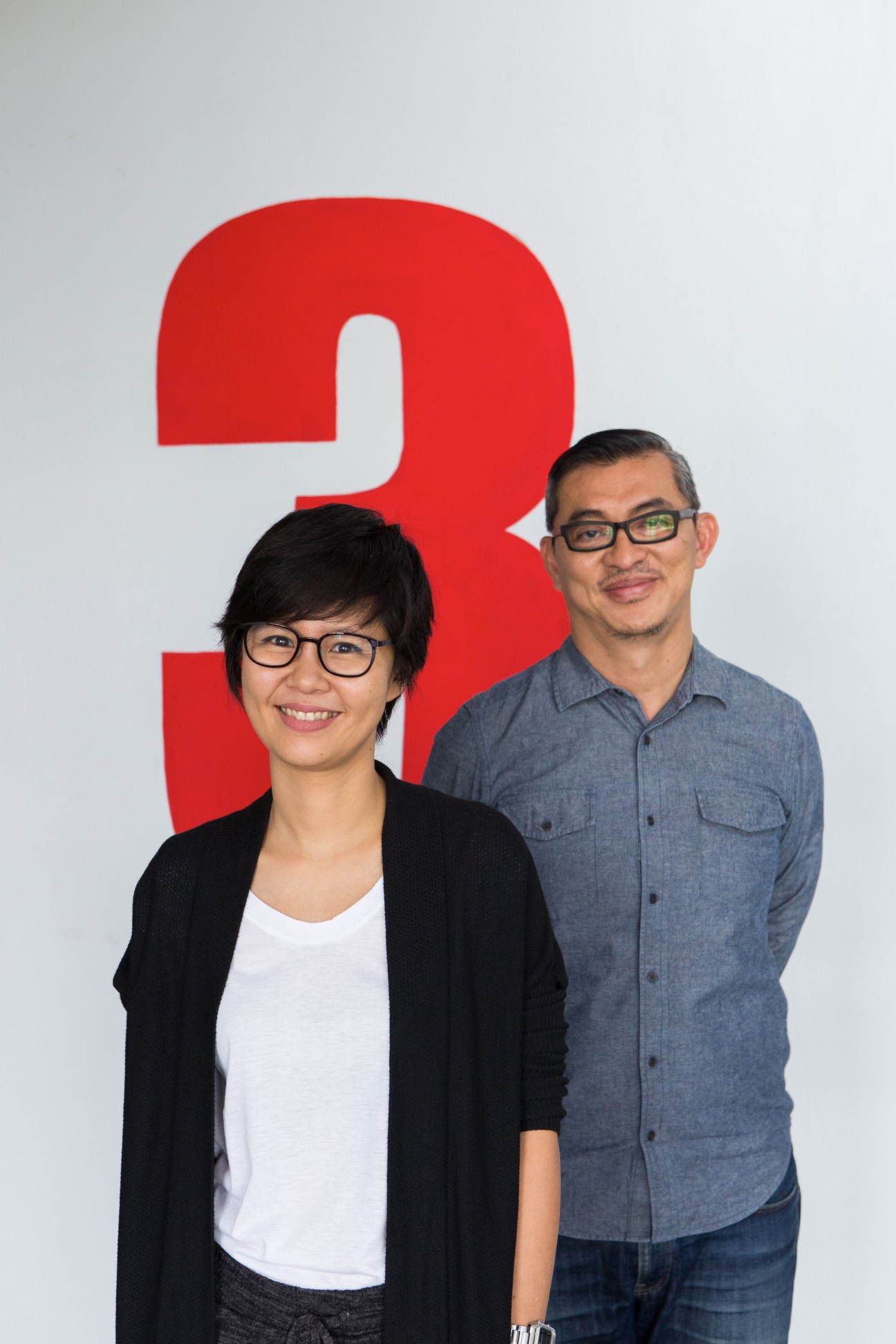 Two people wearing glasses standing in front of a large red number three on a white wall