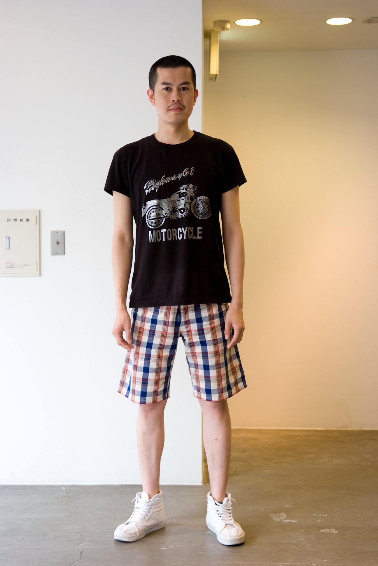 Yoshikazu Miyazaki, an A.P.C. worker, stands in a minimalist interior wearing a black motorcycle graphic t-shirt and plaid shorts.