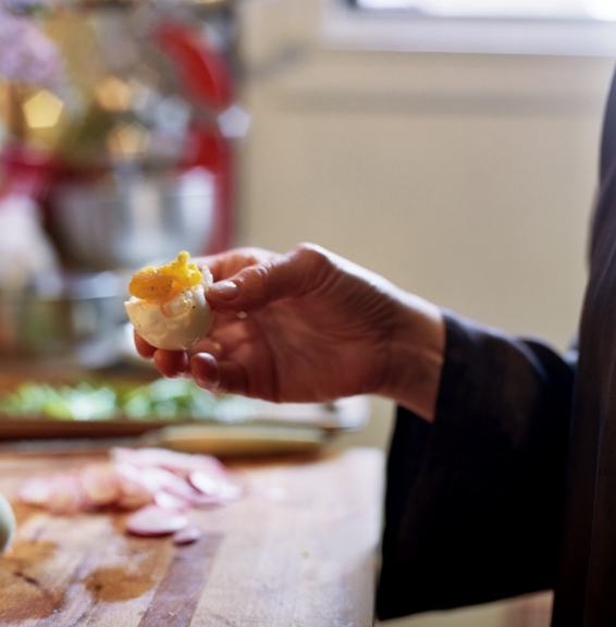 A hand holding a boiled egg with golden yolk visible, with kitchen ingredients blurred in background.