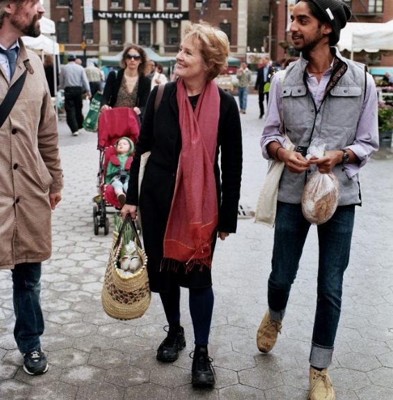 Alice Waters walks through a farmers' market with two companions, carrying woven baskets filled with fresh produce.