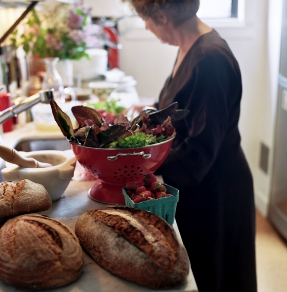 Alice Waters prepares fresh greens in a red colander in her kitchen with bread and produce on the counter.